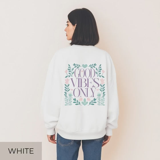 Eine Frau steht mit dem Rücken zur Kamera und trägt einen Oversized-Sweater. Auf der Rückseite befindet sich ein großes florales Design in Grün, Rosa und Flieder, das den Schriftzug „Good vibes only“ einrahmt. Sie trägt eine helle Jeans und steht in einem gemütlichen, hell eingerichteten Raum mit Pflanzen und weichen Textilien im Hintergrund.